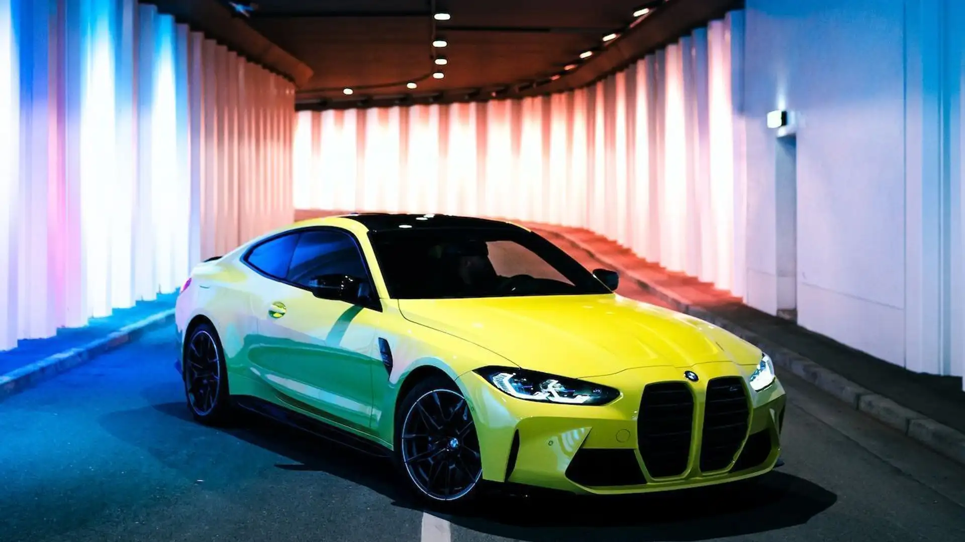 A yellow BMW M4 Competition stands at an angle in an illuminated underpass.